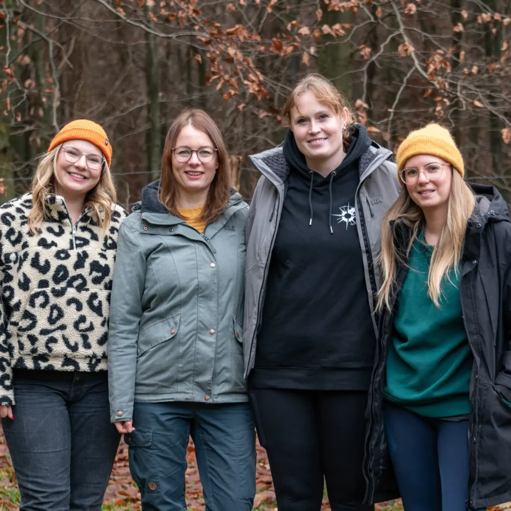 Vier Frauen im Wald, lächelnd, in Freizeitkleidung und warmen Mützen.