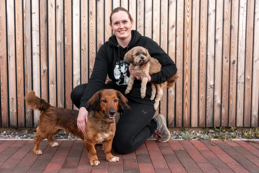 Frau kniet mit zwei Hunden auf einem Steinweg vor einer Holzlattenwand.